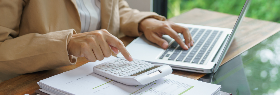 Man sieht eine Frau, die mit Taschenrechner und Unterlagen am Laptop arbeitet.